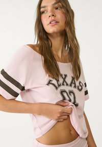 Roze T-shirt met korte mouwen, met een zwarte print "TEXAS 02" en zwarte strepen op de mouwen. Zachte stof en een relaxte pasvorm.