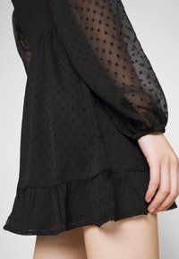Black dress featuring sheer puffed sleeves, textured fabric with small polka dots, and a ruffled hemline.