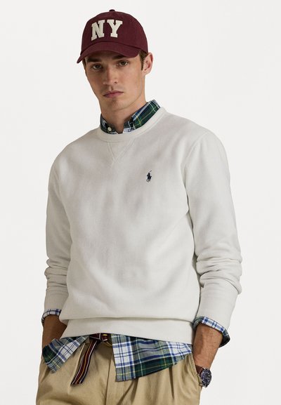Jonge man met een bordeauxrode NY baseballpet, een witte trui over een geruit overhemd, beige broek en een polshorloge, staand met handen in de zakken.