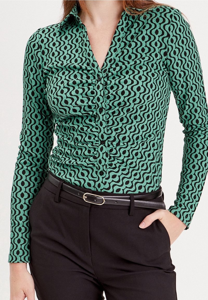 Femme portant une chemise verte à motifs à manches longues rentrée dans un pantalon noir avec une slim ceinture noire, main dans la poche, sur fond blanc.