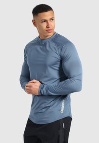 Langärmliges Sportshirt in gedämpftem Blau. Glattes, leichtes Material mit Raglanärmeln und abgerundetem Saum. Mit weißem Logo an der Seite.