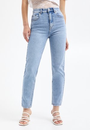 Kvinde iført lyseblå jeans med høj talje og hvide, remsandaletter med hæl, står mod en hvid baggrund.