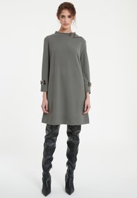 Gray long-sleeve dress with a high neckline and a decorative buckle detail on the shoulder, paired with fitted black leather thigh-high boots.