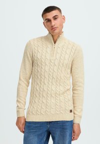 Cremfarbener Zopfmuster-Pullover mit hohem Kragen und halbem Reißverschluss. Gerippte Bündchen und Saum, aus weichem, strukturiertem Stoff gefertigt.