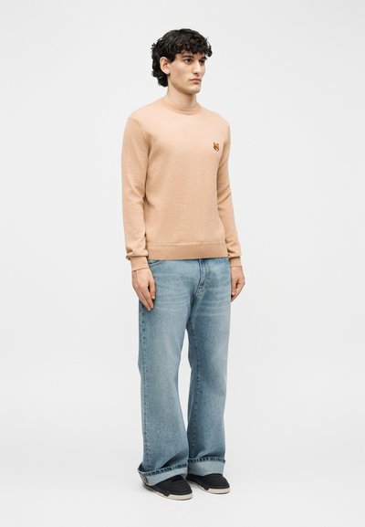 Homme debout portant un pull marron clair avec un emblème de renard, un jean large bleu clair et des baskets noires sur un fond blanc uni.