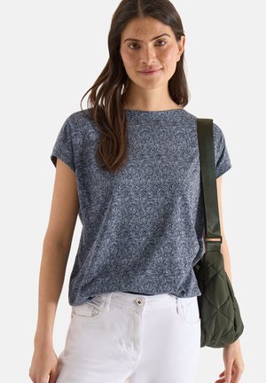 Ung kvinde med langt brunt hår iført en blå t-shirt med blomsterprint, hvide bukser og bærende på en grøn quiltet skuldertaske.