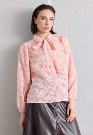 Žena oblečená v priesvitnej ružovej čipkovanej blúzke s kvetinovým vzorom, s golierom na mašľu a dlhými rukávmi, v kombinácii s čiernou koženou sukňou na gombíky.