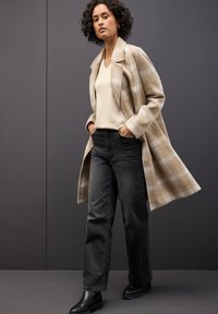 Beige kostkovaný vlněný kabát přes světlý svetr a černé rovné džíny. Černé kotníkové boty s hladkým povrchem dotvářejí celý outfit.