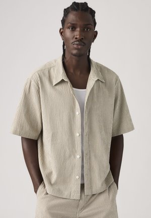 Homme aux cheveux tressés portant une chemise à manches courtes beige à carreaux et un pantalon assorti par-dessus un débardeur blanc, debout les mains dans les poches.