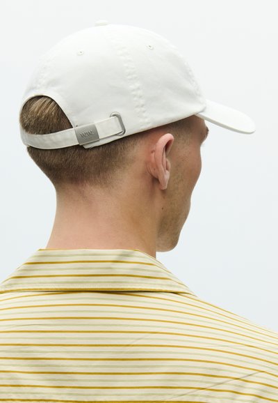Λευκό βαμβακερό καπέλο μπέιζμπολ με καμπυλωτό γείσο και ρυθμιζόμενο πίσω λουράκι με μεταλλικό κούμπωμα. Το μοντέλο φοράει κίτρινο ριγέ πουκάμισο.