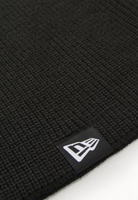 Black knitted beanie with textured pattern, featuring a small black and white logo tag on the edge. Material appears soft and flexible.
