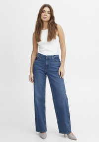 Jeans de tiro alto y pierna ancha de color azul con una textura de denim suave, complementados con una blusa sin mangas blanca. Los zapatos son de un gris claro con punta afilada.