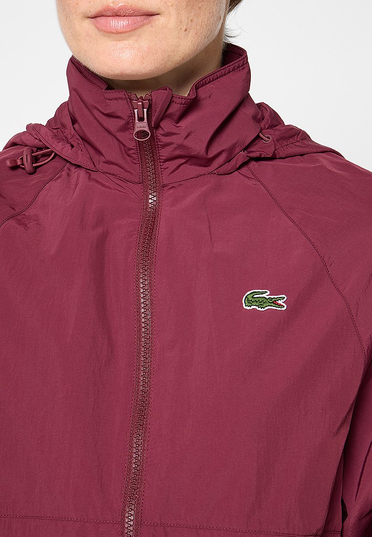 Bordeaux kleurige lichtgewicht jas met een ritssluiting aan de voorkant, hoge kraag, verstelbare trekkoord capuchon en geborduurd krokodillenlogo op de borst.