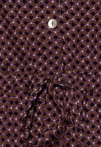 Tessuto marrone a motivo con forme geometriche, caratterizzato da un bottone bianco e una cintura di tessuto legata. La texture appare liscia e leggera.