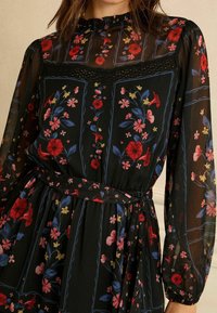 Fekete virágos ruha hosszú, áttetsző ujjakkal. Vörös és kék virágmintákkal, fodros nyakkivágással, övvel és redőzött derékrésszel.