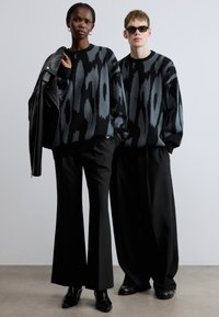 Črni in sivo vzorčasti puloverji z dolgimi rokavi, združeni z visokim pasom črnih hlač. En model drži usnjeno jakno.