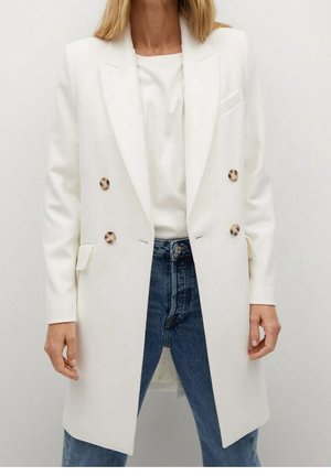 Frau trägt einen langen weißen Doppelreiher-Blazer mit braunen Knöpfen, weißem Oberteil und blauen Jeans vor einem schlichten Hintergrund.