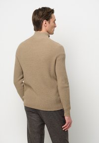 Beige Rippstrickpullover mit hohem Kragen, tailliertem Schnitt und langen Ärmeln, präsentiert vor einem schlichten Hintergrund mit minimalen Details.