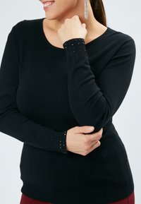 Schwarzer Pullover mit langen Ärmeln und rundem Ausschnitt. Rippbündchen verziert mit kleinen Perlen, glatte Textur und taillierte Passform.