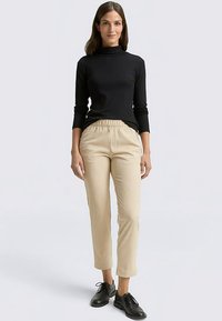 Zwarte coltrui met lange mouwen gecombineerd met beige geribbelde broek. De broek heeft een elastische tailleband en het model draagt zwarte schoenen.