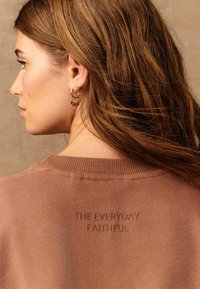 Vrouw met lang bruin haar, die gouden oorbellen en een bruine trui draagt met de tekst "THE EVERYDAY FAITHFUL" geborduurd op de achterkant.
