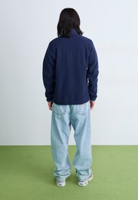 Navy fleece jacket with a high collar, worn over light blue baggy jeans. The fabric has a soft texture, featuring a minimalistic design.