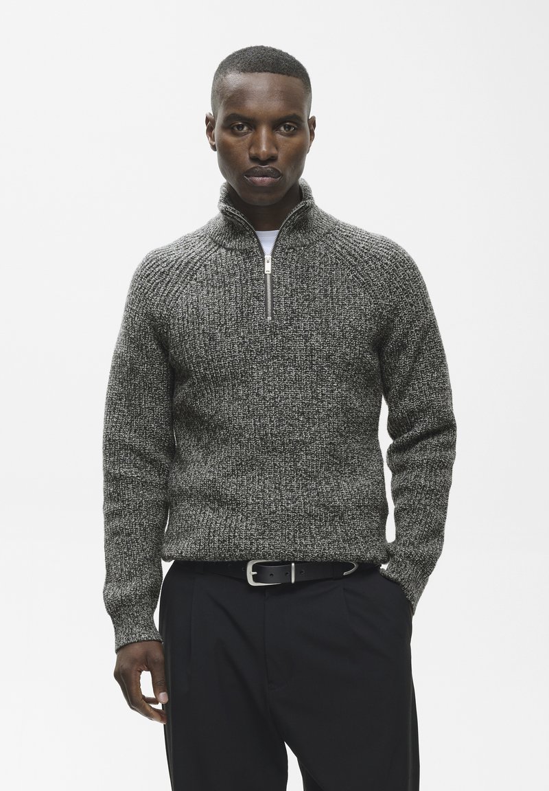 Homme portant un pull en maille gris à fermeture éclair et un pantalon noir avec une ceinture noire, debout devant un fond blanc uni.
