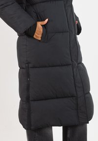 Person iført en lang sort puffjakke med lynlåsdetaljer på siden og hænderne i forlommerne, sammen med mørk denim jeans.