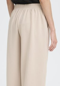 Pantaloni beige a gamba larga con vita elastica, caratterizzati da una texture liscia e tasche laterali. Il design è minimalista ed elegante.