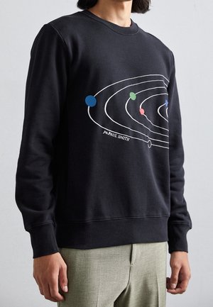 Person iført sort sweatshirt med farverigt, stiliseret solsystemdesign og teksten "ps PAUL SMITH", kombineret med lysegrå bukser.