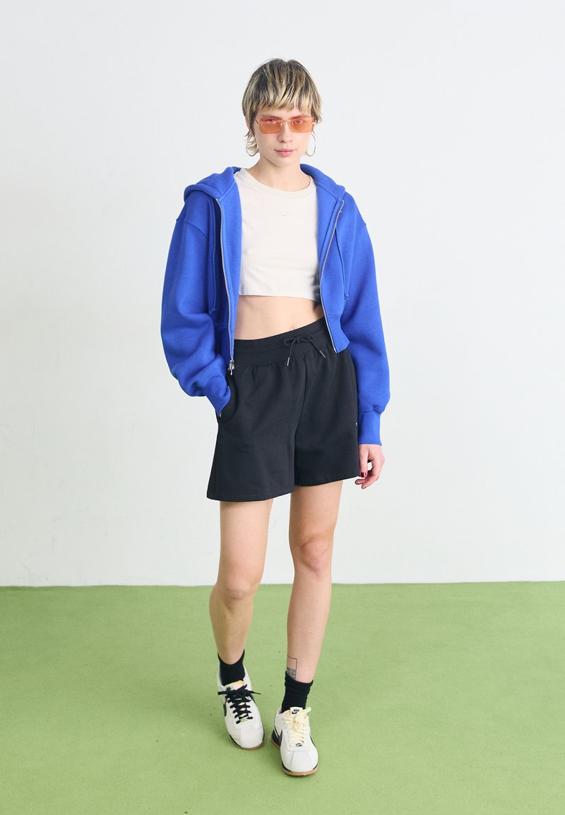 Sudadera corta azul, camiseta blanca, pantalones cortos negros, calcetines negros y zapatillas bicolor. Modelo con gafas de sol naranjas, de pie sobre un suelo verde.