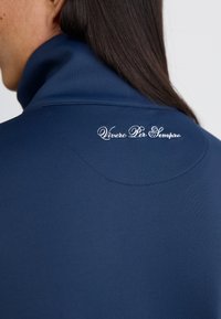 Temno modra majica s visokim ovratnikom, izdelana iz gladkega materiala. Na hrbtni strani, blizu ovratnika, je izšito besedilo "Vivere Per Sempre".