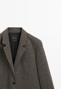 Blazer en tweed gris foncé avec revers crantés, un seul bouton noir et une poche poitrine passepoilée, présenté sur fond blanc.