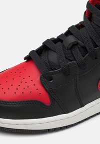 Black leather sneaker with red accents and perforated toe. Features black laces, a white midsole, and a smooth texture.