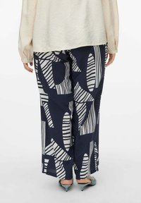 Pantaloni a fantasia navale con forme astratte bianche e strisce, realizzati in tessuto leggero, abbinati a una blusa crema chiaro e tacchi color teal.