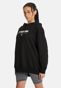 Sudadera negra con bolsillo canguro frontal, cordones y gráficas blancas en negrita. Tejido suave, corte holgado y bajo trasero extendido.