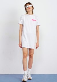 Hvit kortermet kjole med en rosa logo på brystet, kombinert med hvite sporty solbriller og grå sneakers, stående mot en ensfarget bakgrunn.