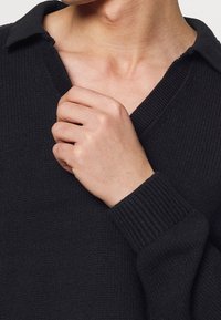 Maglione nero lavorato a maglia con scollatura a V e polsini a costine, caratterizzato da un tessuto texturizzato. La persona sistema il colletto con una mano.