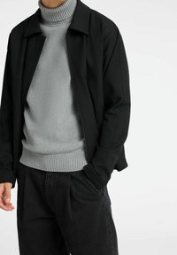 Chaqueta negra con cremallera sobre un suéter de cuello alto de canalé gris claro, combinada con pantalones oscuros. Confeccionada en tela lisa y corte relajado.
