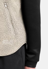 Gilet in pile di un beige chiaro con superficie testurizzata, dotato di zip laterale e maniche lunghe nere sotto. Logo Jack Wolfskin visibile.