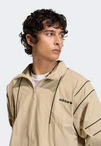 Μπεζ μπουφάν με φερμουάρ και υφή, με μαύρες λωρίδες ως διακριτικά στοιχεία. Λογότυπο στο στήθος και σχέδιο με ψηλό γιακά.