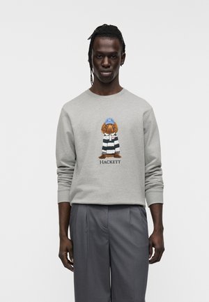 Grå sweatshirt med lange ærmer med en tegneseriehund iført en stribet trøje, med ordet "HACKETT" nedenunder. Modellen bærer grå bukser.