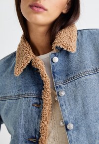 Džínová bunda s béžovým, texturovaným sherpa límcem. Obsahuje kovové knoflíky a má strukturovaný střih. Nosená přes pletený světle zbarvený top.
