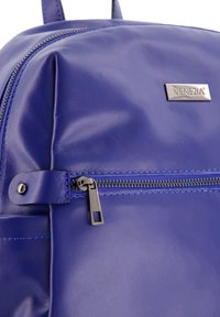 Section de sac à dos en simili cuir bleu avec un logo argenté, une fermeture éclair visible et des coutures détaillées le long des coutures.