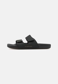 Sandale noire avec deux brides réglables en Velcro, semelle en caoutchouc et détail de logo en relief. Fabriquée en matériau semblable à du daim avec un design plat.