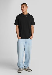 T-shirt nera a collo rotondo, vestibilità comoda, abbinata a jeans azzurri chiari. Modello che indossa sneaker grigie con accenti di blu navy e dettagli bianchi.