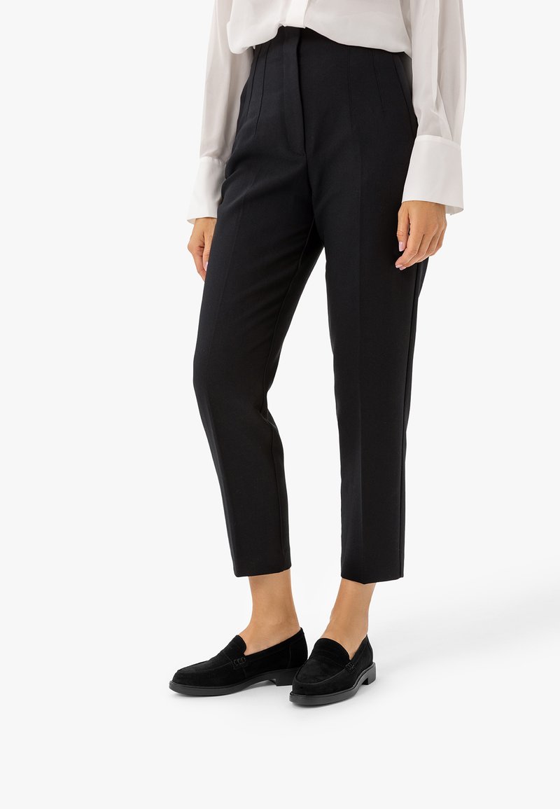 Femme portant un pantalon noir tailleur longueur cheville, une chemise blanche à manches longues et des mocassins noirs sans lacets, debout sur un fond blanc.