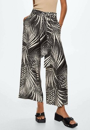 Pantalon à jambes larges imprimé feuilles noir et blanc avec ceinture assortie à la taille, porté avec des sandales à talons noires et un haut beige sans manches.