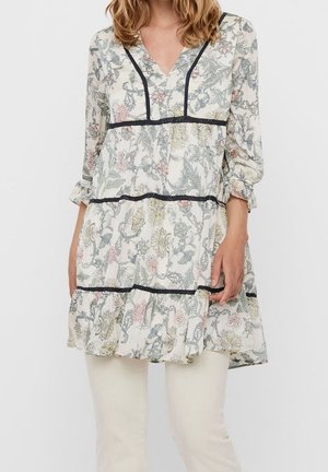 Femme portant une robe tunique à manches longues à motifs floraux avec des bordures noires, associée à un pantalon clair, sur un fond uni.