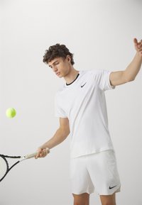 Hvit Nike treningst-skjorte med svarte detaljer, kombinert med hvite shorts. Personen holder et tennisracket, med en gul tennisball i bevegelse.